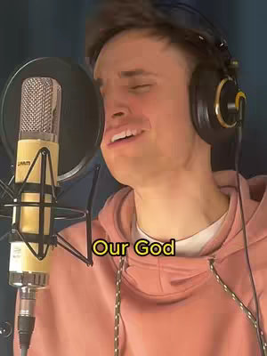 A man in a pink hoodie sings into a microphone with a pop filter. There's an overlay with the text "Our God". The background is a dark blue curtain. He wears headphones.