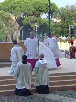 A wide shot capturing a religious ceremony outside, with a focus on a group of people near stairs, and a tall green tree on the left. Several individuals in white robes with different colored accents are central, suggesting a procession with religious figures. The background includes a tree-filled area, and a series of canopy tents in the distance.