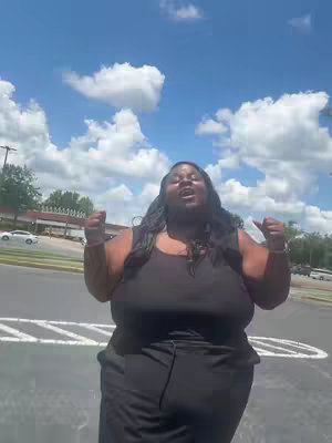 A woman stands outdoors in a parking lot, arms raised with fists clenched near her shoulders. Behind her is a bright blue sky filled with fluffy white clouds. She has long, dark hair and is wearing a black ensemble. The setting appears to be a sunny day.