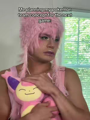 A person with pale skin and expressive makeup is centered in the frame. They wear a pink, feathered headpiece and a matching top. They are holding a pink and yellow plush toy of a Pokémon. The background includes a window with blinds.