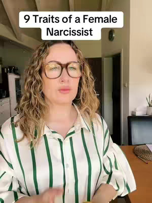 A medium shot of a woman with curly blonde hair, wearing large-framed glasses and a vertically striped blouse. The text overlay reads '9 Traits of a Female Narcissist'. The background is a room interior.