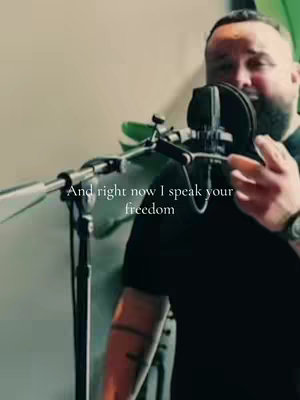 A man wearing a black t-shirt stands in front of a microphone, likely in a recording studio. Text overlayed in white reads, 'And right now I speak your freedom.' The image captures a moment during a worship service or musical performance.