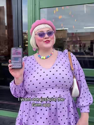 A woman with blonde hair and purple sunglasses holds a phone showing an app screen. She wears a pink beret, a beaded necklace, and a purple dress with black heart patterns. She smiles and stands in front of a green door with a window.