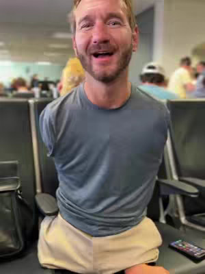 A medium shot of a man sitting in an airport terminal, looking at the camera while smiling. He has short brown hair and a beard. He's wearing a gray t-shirt and beige shorts. Background includes rows of chairs, and the blurry figures of other people, suggesting an airport environment. There is a black bag on the left side of the image and a phone right up in front of his body.