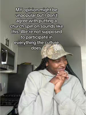 A woman with long black braids is shown in a kitchen setting, wearing a camouflage baseball cap and a gray hoodie. She is positioned in front of a microwave and cabinets. Overlayed text is present, placed above her head. Her expression is one of contemplation, with hands clasped near her face, and her lips slightly pursed.