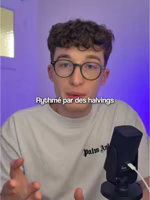 A young man with glasses and curly hair is centered in the frame. He is wearing a white t-shirt, and is speaking, his hands gesturing. Below him, overlaid text reads: 'Rythmé par des halvings'. In the foreground, a microphone is positioned to the right, and the background is a gradient of soft purple and a yellow light on the left.