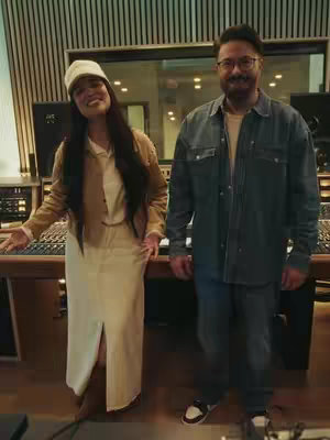 A medium shot taken in a recording studio shows two people, a woman with a white hat and a man with glasses, smiling and looking at at the camera. They are standing in front of a sound mixing board.