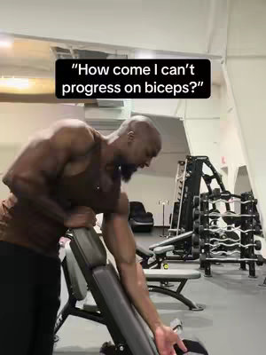 An individual is captured mid-exercise in a gym setting. The person, with a muscular physique and a bald head, is positioned at the left, partially bent over a preacher bench to perform a bicep curl. The image is dominated by shades of brown and gray, creating a subdued and focused ambiance. A text overlay in a black box on top reads, ''How come I can't progress on biceps?'' The setting includes gym equipment, such as weight racks and benches, in the background. The lighting highlights the person and equipment.