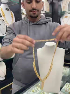 A man in a gray hoodie is holding a gold necklace in front of a display case. The jewelry case is illuminated, with gold necklaces also on display behind the man as well .