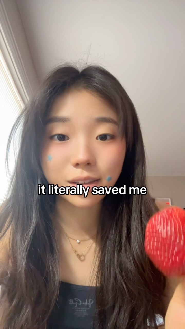 A young Asian woman with long dark hair is centered in the frame, looking towards the camera with a soft expression. She appears to be in a well-lit room. The woman's skin has a light make-up, with a touch of blush. She has two small blue circular face stickers on her cheeks. The woman is holding a brightly colored, red, spherical object in her right hand. Above the woman is a text layer.