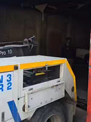 The image shows a large, white tow truck with yellow accents parked inside a dark garage or storage space. The tow truck has open compartments revealing some of its inner workings. A person, their features obscured by the shadows, stands in the background. The overall lighting is dim, giving the setting a somewhat enclosed and shadowy feel.