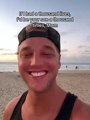 A man is smiling at the camera on a beach. He is wearing a black cap and tank top. In the background, the ocean and a few people are visible.