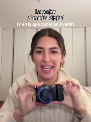 A woman in a beige sweater holds a black camera in front of her, smiling at the viewer. Behind her is a neutral-toned indoor setting with large white doors. Text overlays the image, and her posture suggests she is presenting or reviewing the camera.
