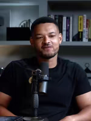 A medium shot features a man, centrally positioned, with short, dark hair and a closely trimmed beard. He's wearing a black t-shirt and facing the camera, with a microphone in front of him. Behind him, a bookshelf holds various books, and a dark cabinet sits to his left.
