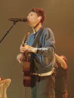 A live performance photo of a man singing and playing an acoustic guitar on a stage. He is wearing a blue sweater and white shirt. The background is a dimly lit stage with other musicians slightly visible. Text overlay 'OH LAMB OF GOD' in white.