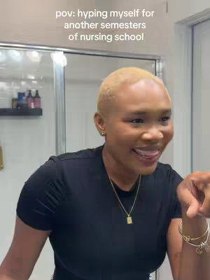 A woman with blonde hair stands in a bathroom, looking in the mirror and pointing. The upper body is visible, wearing a black top and gold necklace. Behind her is a partially visible, slightly blurred background with a shower area.