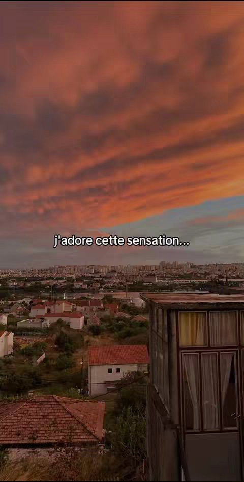 The image showcases a sunset view over a city, with muted orange and pink hues dominating the sky. Below, a cityscape unfolds, and a house, with an attached balcony where someone has placed a curtain in the windows on the rights, anchors the foreground.