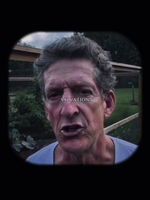 A man with graying hair, wearing a blue shirt, looks directly at the camera against a backdrop of greenery. A text overlay displays the word 'SALVATION'. The image is framed within a blurred oval vignette, creating a focus on the man's face.