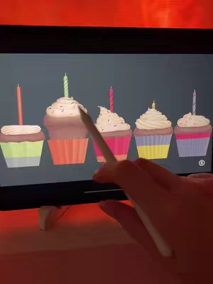 The cover image shows an iPad screen displaying five cartoon cupcakes, each with a candle. A hand holding a stylus is interacting with the image, specifically pointing at one of the cupcakes. The background is a blurry orange color.
