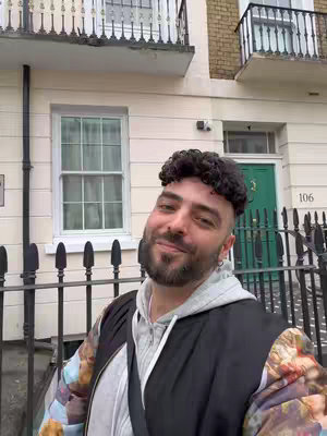 A medium shot of a man with curly hair and a beard standing in front of a building with a green door and white facade. He is wearing a black jacket over a gray hoodie. A black fence is in the foreground. The building has white-framed windows and a small sign to the left of the door.