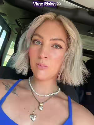A selfie-style shot featuring a woman with light blonde, short hair posing inside a vehicle. She wears a blue top and has a double-layered necklace with a heart pendant. The hook 'Virgo Rising 👀' appears at the top of the frame. The background is blurry, but car interior and some greenery are visible.