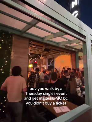 An outdoor scene at dusk. The camera faces a lit bar entrance, where a crowd of people inside are visible through the large windows. A person stands in the foreground, facing away. The image is bathed in warm light from inside the bar, contrasted with the twilight outside.