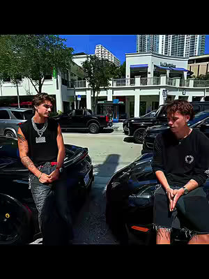 Two young men are positioned near black cars, leaning on them. They are on a street in front of store fronts and a blue sky. One man is wearing a sleeveless shirt and heavy jewelry. The other is on the right in a long-sleeve shirt. The composition is symmetrical with the cars and men creating a visual balance.