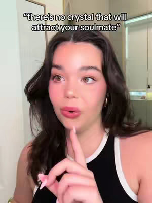 The cover image features a young woman with dark hair and a black top, her face close to the camera. She's looking towards the side with a slightly concerned expression and a raised index finger, as if making a point. The background is neutral, with a glimpse of what appears to be a white wall in the background. Text overlayed on the image reads 'there's no crystal that will attract your soulmate.'