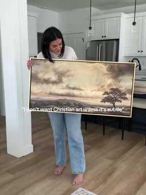 A woman is standing barefoot in a brightly lit room holding an art piece. The artwork is rectangular, with a gold frame and contains a landscape painting. The painting reflects a subtle sepia tone and features a tree on the right side and a cloudy sky. The woman is smiling, her hair dark around her shoulders, and she is wearing a white shirt and light-wash jeans. A sticker is at the bottom of the image. In the background, there's a kitchen, white cabinets, and a stainless steel refrigerator.