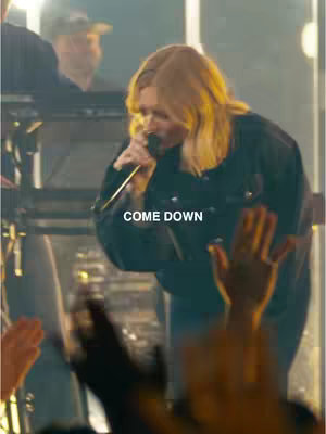 A woman with blonde hair singing into a microphone, with a crowd's hands raised in the foreground. The background is a stage with lighting.