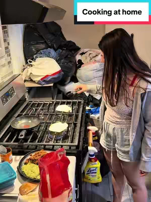 A woman stands in a kitchen, facing the stovetop. She is wearing a light-colored tank top and shorts, and has long, dark hair. On the stove, there are several items in pots and pans. The background is cluttered with bags and laundry. A text overlay at the top of the screen reads "Cooking at home."