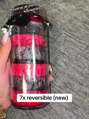 A hand holding a cylindrical, glass jar filled with alternating layers of black and red wax or material. The jar is partially obscured by a black covering at the top. Below, a white box displays the text '7x reversible (new)'. The background is a textured gray surface.