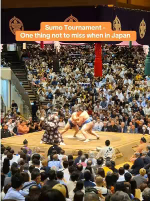 An image taken inside a sumo wrestling arena, filled with a massive crowd observing two sumo wrestlers in the ring. The ring is elevated, with a light-colored clay or sand surface, and the wrestlers are actively engaged in a match. The background details the crowded seating arrangement.