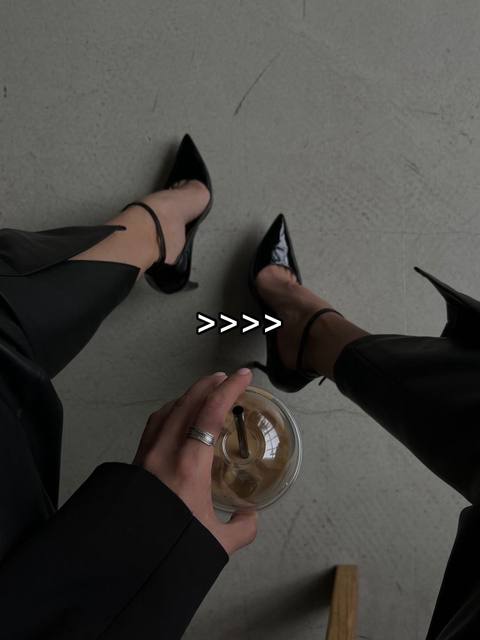 The image shows a top-down view of a person's legs clad in black clothing and heels. The person is holding a cup of coffee. The composition is simple, with the person's legs and the drink being the main focus on a plain gray floor.