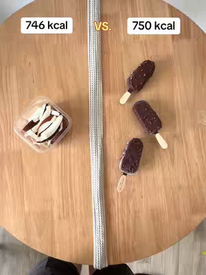 A top-down shot shows a round, wooden table. On the left, a clear plastic container holds several brown and white coconut-based treats. A thin white striped object runs vertically down the center. On the right, three chocolate-covered ice cream bars on a stick are arranged. Text overlays at the top label one group as '746 kcal' and the other as '750 kcal'.