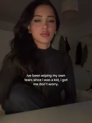 A young woman with long dark hair and fair skin gazes directly into the camera. She wears a black long-sleeve top in a dimly lit room with soft lighting. Text is overlaid in white at the bottom of the frame.