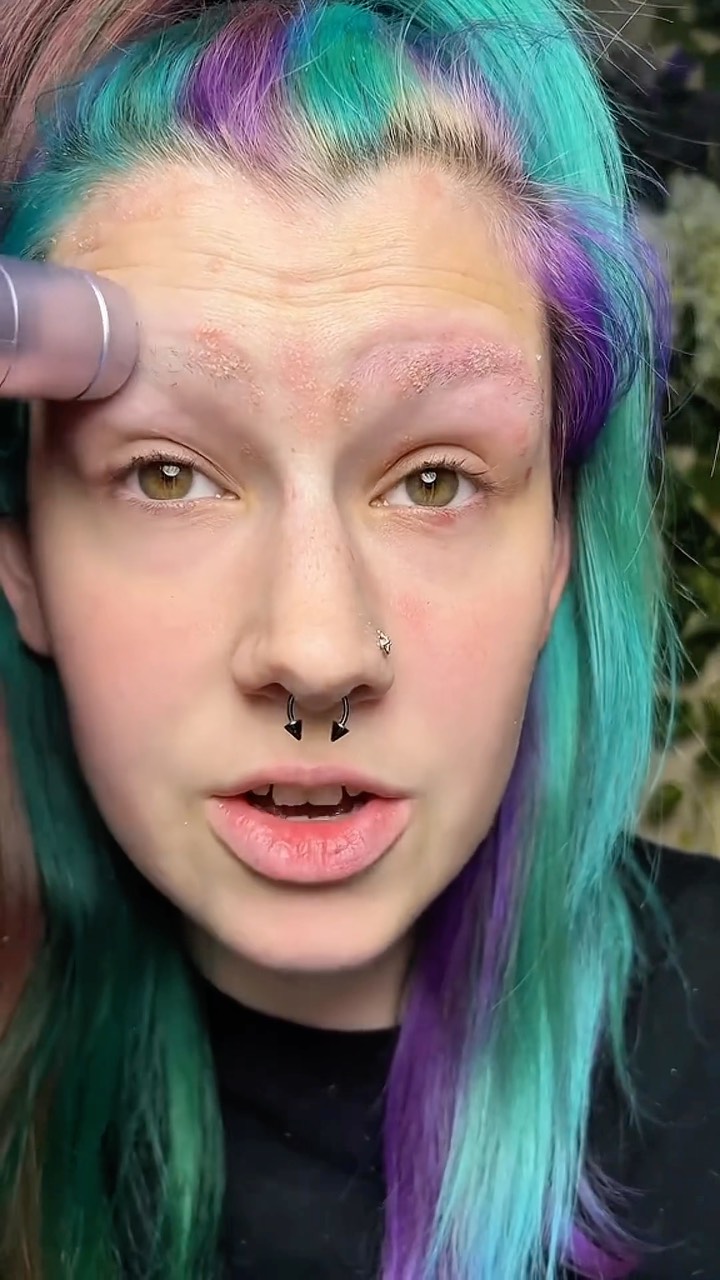 A close-up shot of a person's face with colorful hair and a grooming tool. The person stares at the camera with a neutral expression. There are signs of recent grooming on the eyebrows.