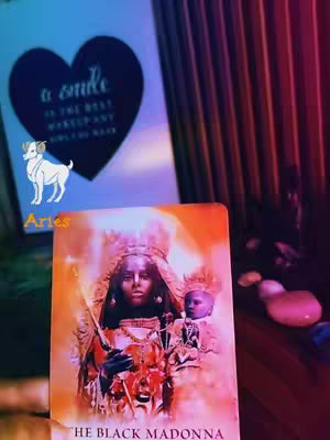 The cover image features a tarot card depicting a woman-like figure with a child, overlaid on a background filled with colorful lights. In the bottom part, the card displays the words: 'THE BLACK MADONNA'. The background includes what seem to be blurred items and overall enhances the spiritual vibe. There are Arabic texts in the image like برج_الحمل (Aries) and a heart shape with the words 'a smile is the best makeup, girt it again' which might be the hook text. There is also an Aries illustration
