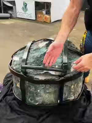 The cover image features a close-up shot of a large, heavy-looking object, possibly a crystal or gemstone, encased in a clear container. The object appears to be a large, irregularly shaped mass with a mottled green and clear appearance, enclosed in a clear container or display. The container rests on a black surface. A person’s arm and hand are visible, interacting with the object, suggesting they are handling or examining it. In the background, there's a glimpse of a shop or area with merchandise, possibly a gem or crystal shop.