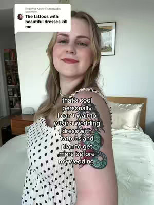 A woman with blonde hair and a tattoo on her upper arm, smiling. The background is a room with a bed and a window.
