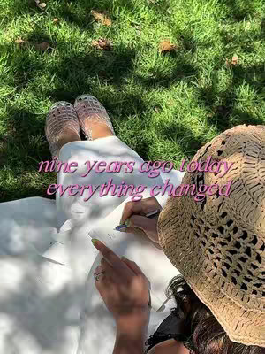 Overhead shot of a person sitting in the grass writing with a pen. They are wearing a straw sun hat and white clothing. The grass is green, and the overall tone is natural and serene. The hook text 'nine years ago today everything changed' is overlaid in pink.