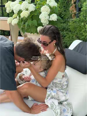 A family moment captured with a woman, a man, and a child on an outdoor sofa with lush greenery in the background. The woman, wearing sunglasses and a white top with a patterned robe, cuddles the child, while the man kisses the child's head. A large vase of white flowers adds a decorative element to the scene, which conveys a sense of intimacy.