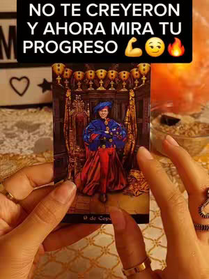 The image shows a person holding a tarot card. The person's hands are in the foreground of the image, with long nails, and various rings can be seen on the fingers. The card itself is of a man seated in a throne, which appears to be inside a church. The background includes blurred elements, such as burning candles that add to the mystical atmosphere.