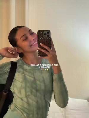A selfie-style video cover image featuring a young woman with dark hair pulled back, smiling at the camera while holding a phone. She is wearing a light green top and a black shoulder bag. Behind her is a room setting with blurred background. The text overlay at the bottom-right corner reads 'DIML AS A PEDIATRIC CNA 4 AM - 8 PM' followed by several emojis.