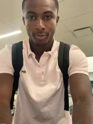 A man wearing a light pink polo shirt and a black backpack looks directly at the camera in what appears to be a well-lit interior space, possibly a hallway. The background is out of focus.