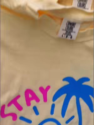 The image shows a folded light yellow tee shirt with a graphic of the word STAY in pink stylized letters with a blue sun with rays and a blue palm tree.