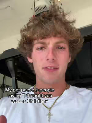 A young man with curly brown hair is centered in a selfie-style shot. He wears a white t-shirt and a gold necklace with a cross pendant. His expression is serious with a slight frown. The background shows a garage-like setting with a ceiling fan, creating an informal, personal feel.