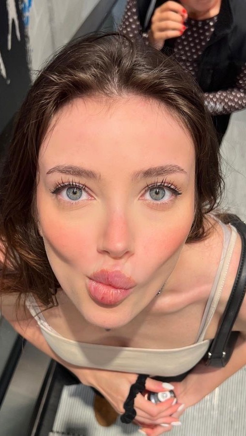 A young woman looking up at the camera, with her lips puckered in a kiss. She has light skin, blushed cheeks, and blue eyes lined with black eyeliner. Her dark brown hair is styled away from her face. She is wearing a light-colored top.