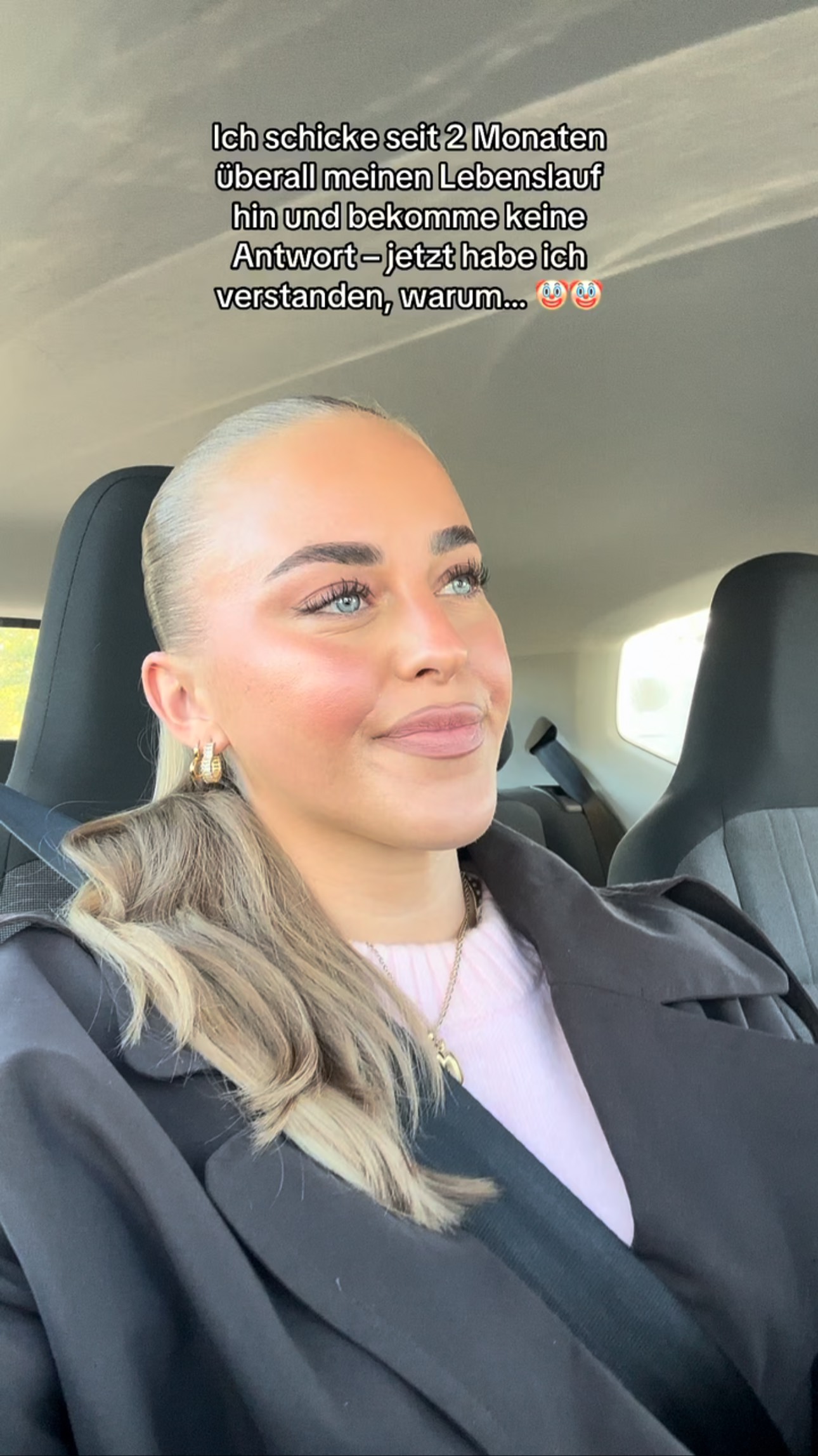 A woman is seated in a car, looking up and to her right with a slight smile. She has blonde hair, a ponytail, and is wearing a black coat over a pink top. The background is the interior of a car, with the seat visible. The overall aesthetic is casual and natural.
