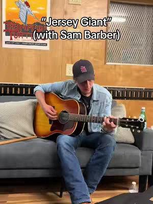 A man sits on a gray couch playing an acoustic guitar. He's wearing a baseball cap, a denim shirt, and jeans. The background includes a partially visible poster, wooden paneling, and a window.